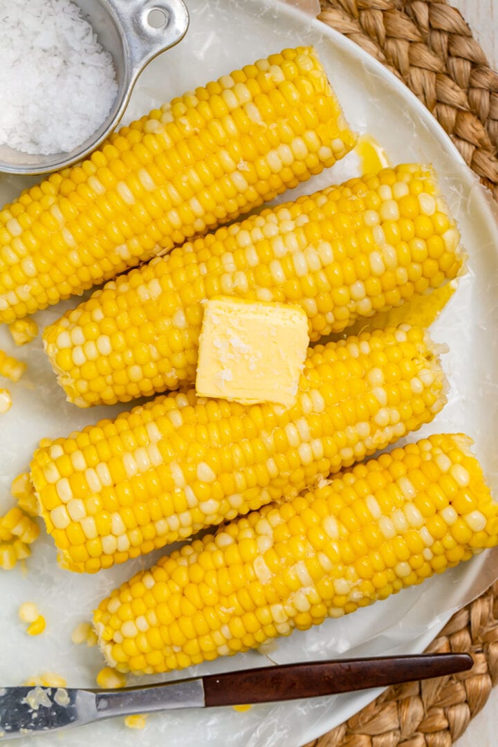 4 ears of boiled corn on the cob laying on a white oval platter on a woven placemat.