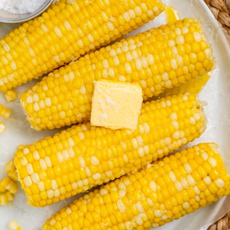 4 ears of boiled corn on the cob laying on a white oval platter on a woven placemat.