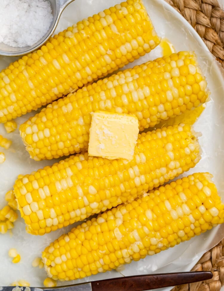 4 ears of boiled corn on the cob laying on a white oval platter on a woven placemat.