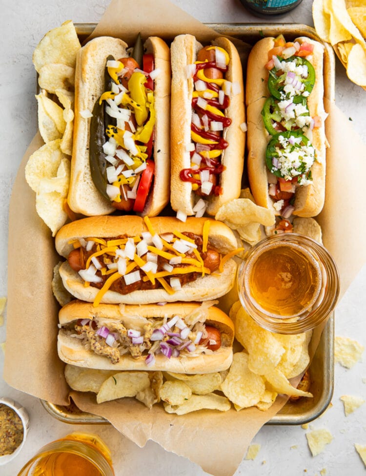 5 air fryer hot dogs in hot dog buns with various toppings arranged on a baking sheet.