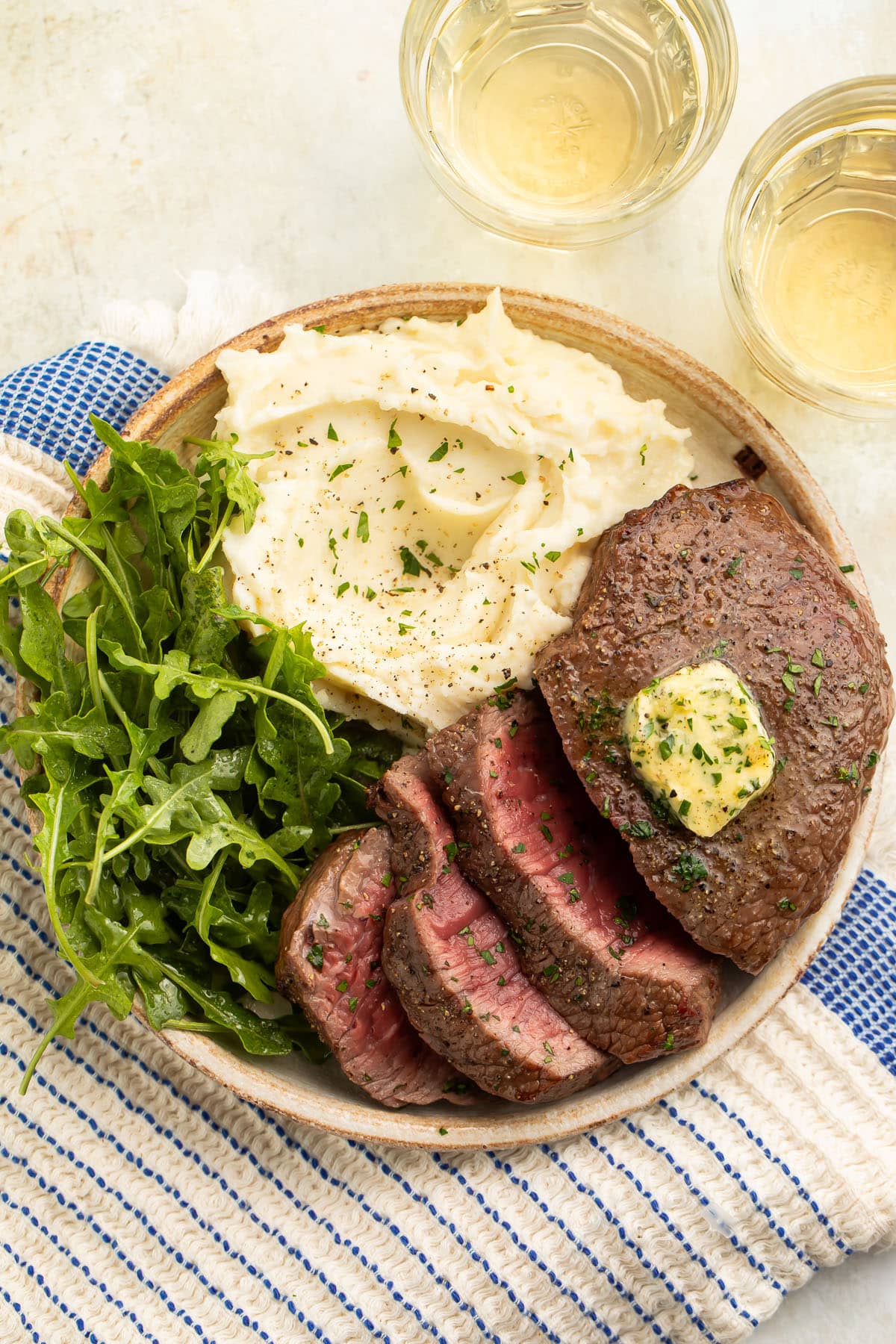 Overhead photo of medium-rare air fryer steak, topped with homemade herb butter, sliced and arranged on a plate with mashed potatoes and a small salad.