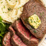 Close-up of medium-rare air fryer steak, topped with homemade herb butter, sliced and arranged on a plate with mashed potatoes and a small salad.