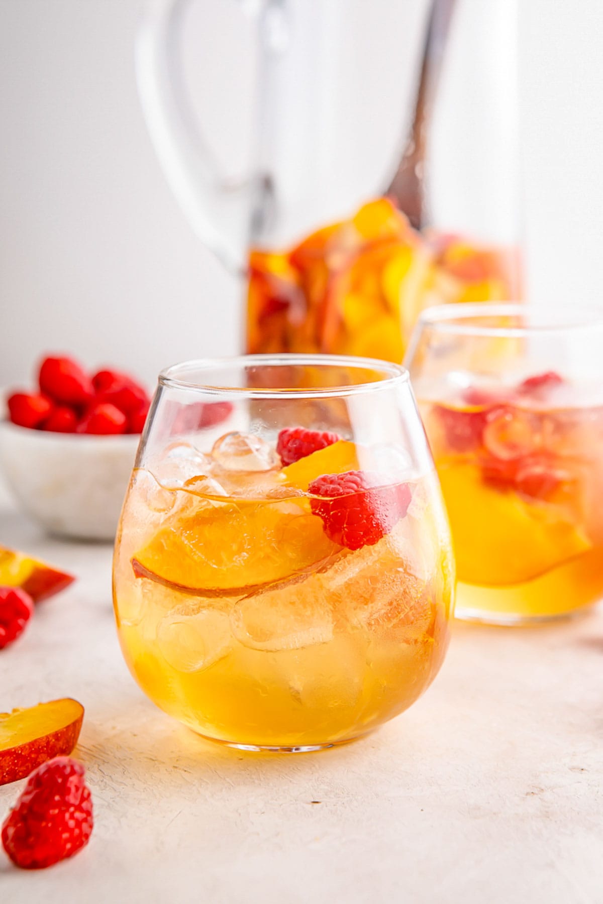 A stemless wine glass of peach white wine sangria on a table in front of a pitcher of sangria.