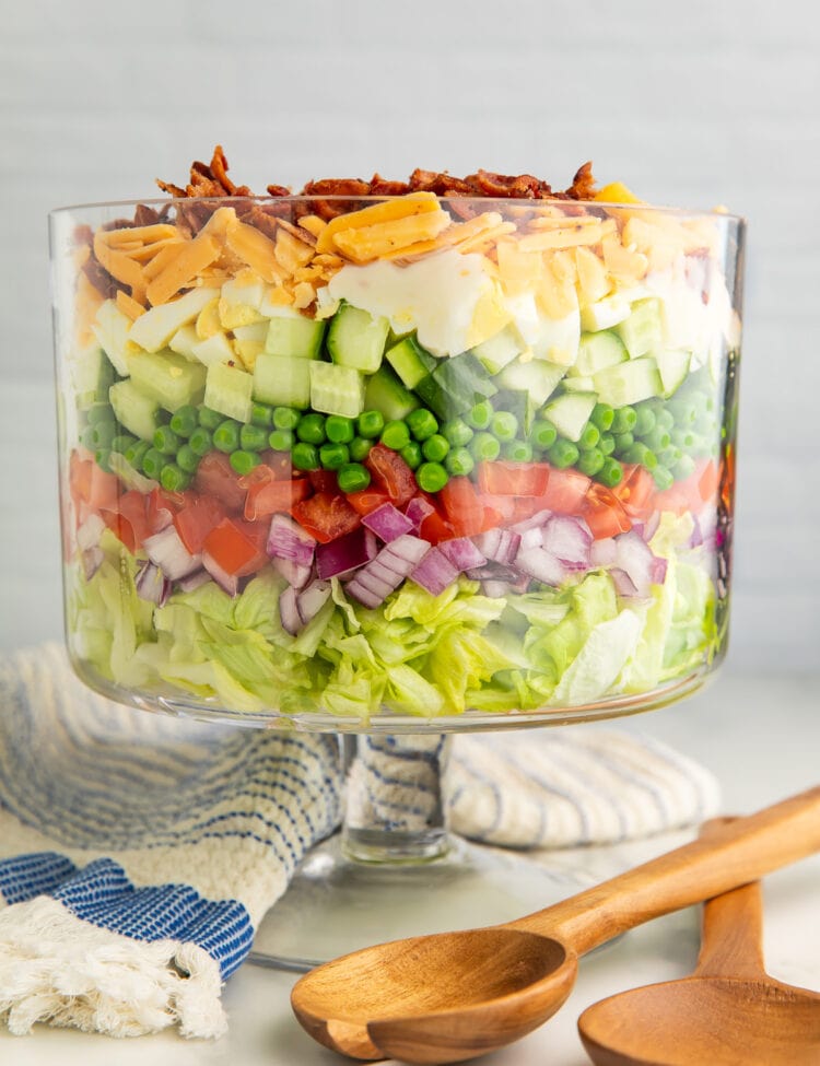 A 7 layer salad in a glass trifle dish with a pedestal base next to wooden utensils and a dish towel.