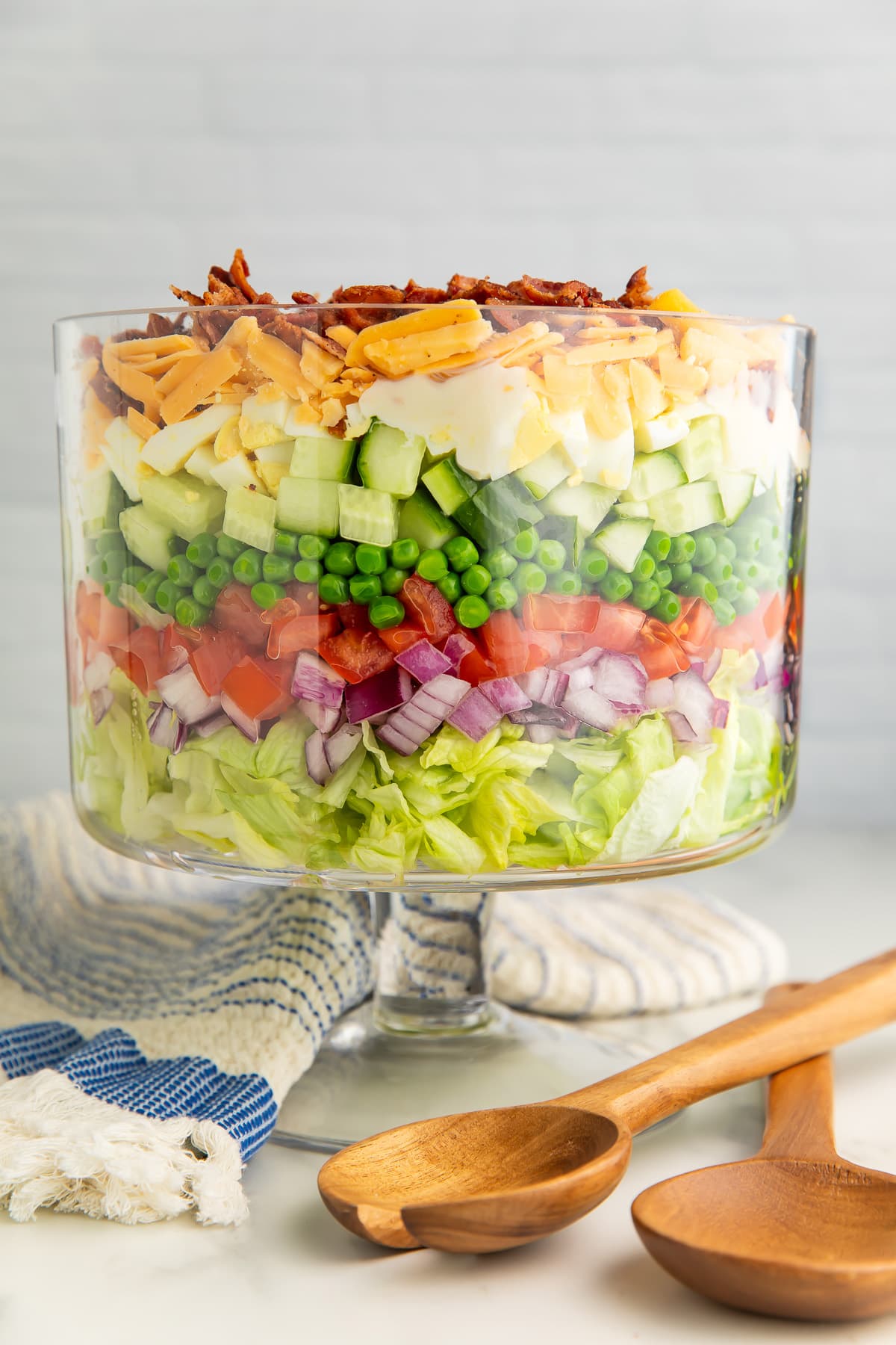A 7 layer salad in a glass trifle dish with a pedestal base next to wooden utensils and a dish towel.