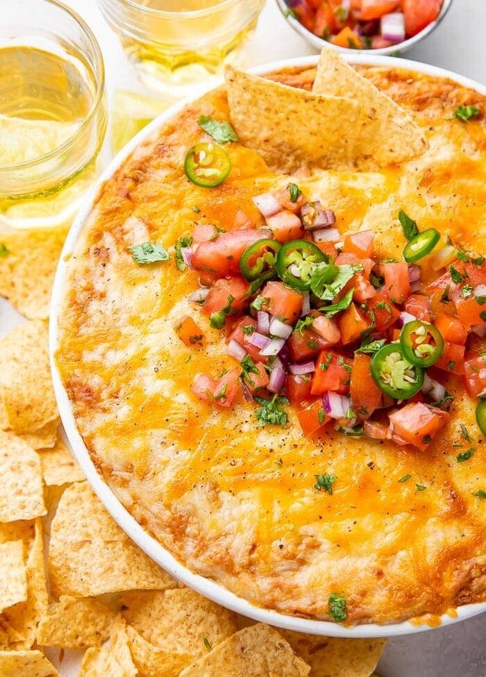 Overhead shot of bean dip in a bowl topped with tomatoes and jalapenos, surrounded by chips and glasses of beer