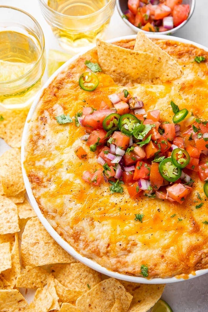 Overhead shot of bean dip in a bowl topped with tomatoes and jalapenos, surrounded by chips and glasses of beer