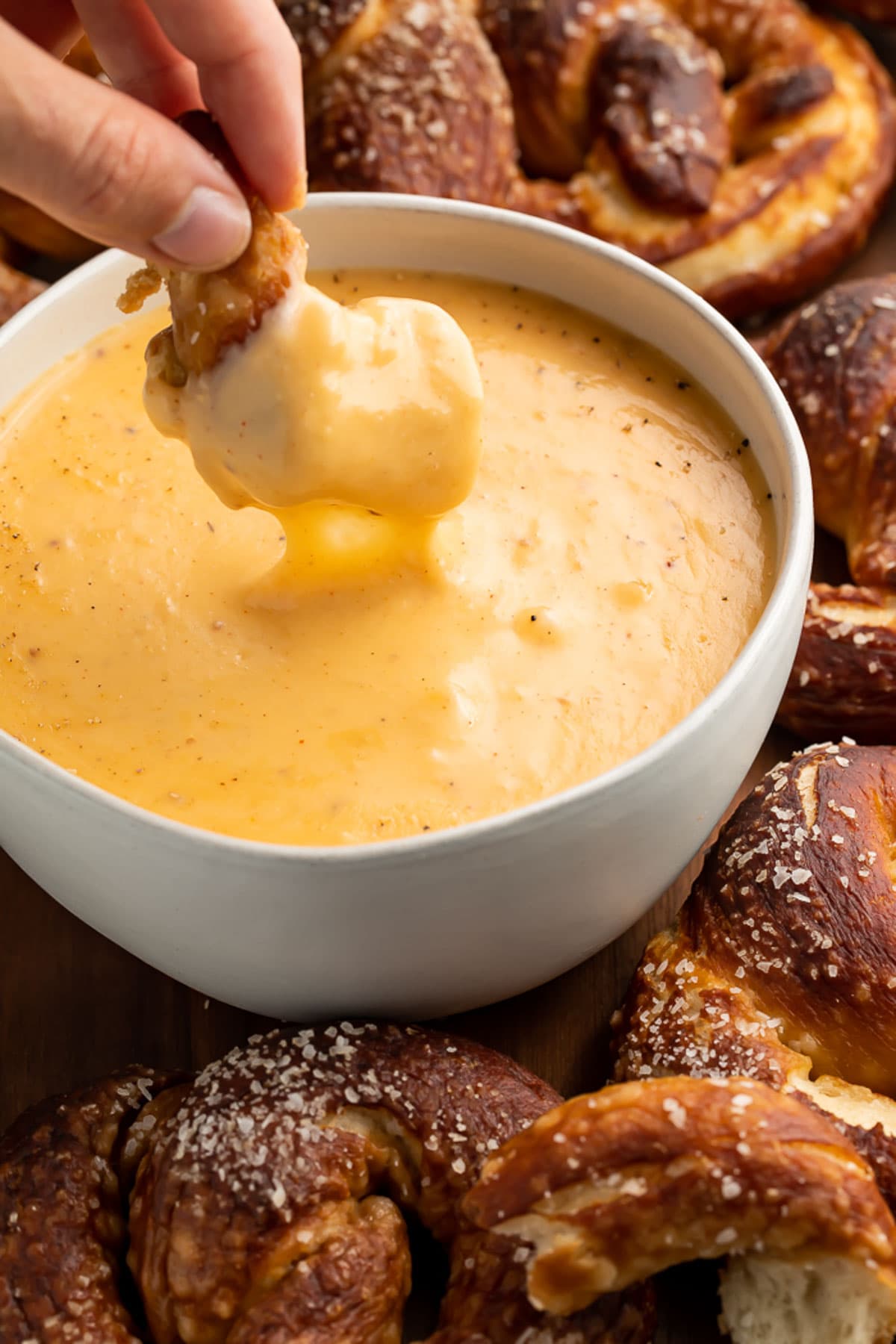 A woman's hand lifting a piece of soft pretzel out of a bowl of beer cheese dip.