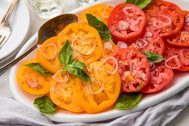 Slices of red and yellow heirloom tomatoes on a serving dish covered in sliced shallots and basil leaves