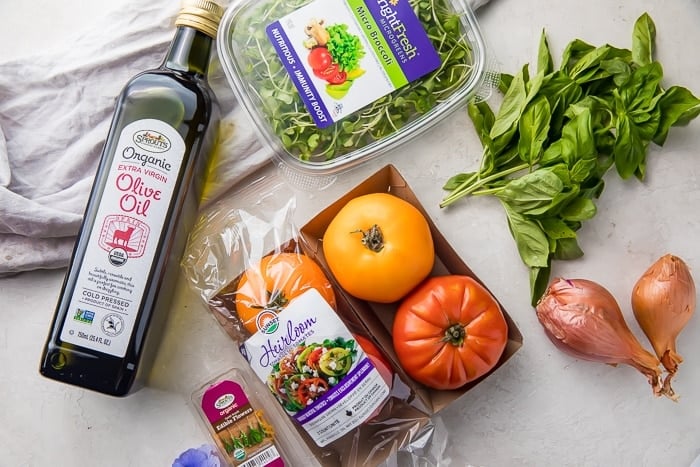 A bottle of olive oil next to a package of differently colored heirloom tomatoes, shallots, basil leaves, and micro broccoli