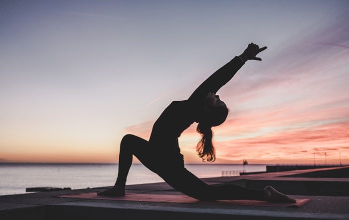 Woman doing yoga by the water in front of a sunset
