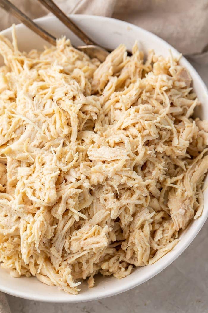 Close-up overhead shot of shredded chicken in a white bowl with 2 metal forks for shredding