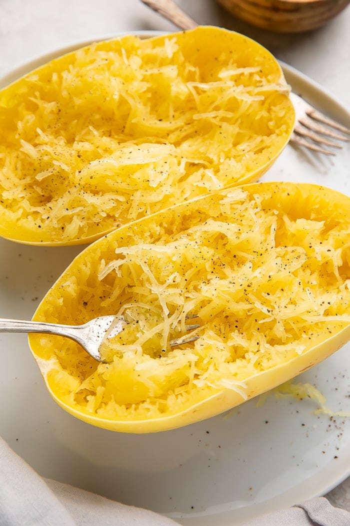 Two halves of cooked spaghetti squash on a white plate with a fork stuck in one half