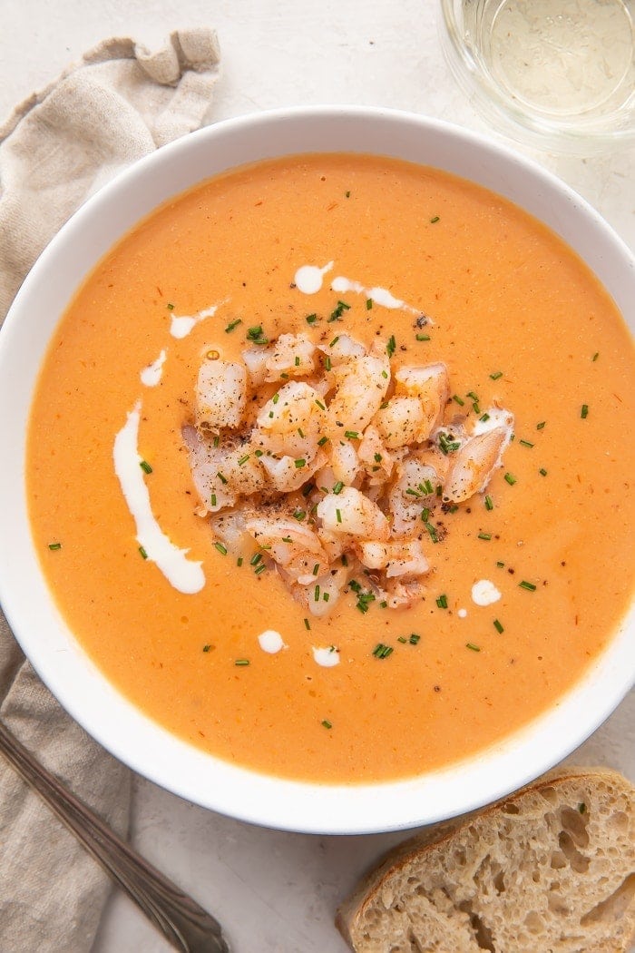 A bowl of shrimp bisque with a pile of shrimp in the center and a circle of cream surrounded by a fork, napkin, and piece of bread