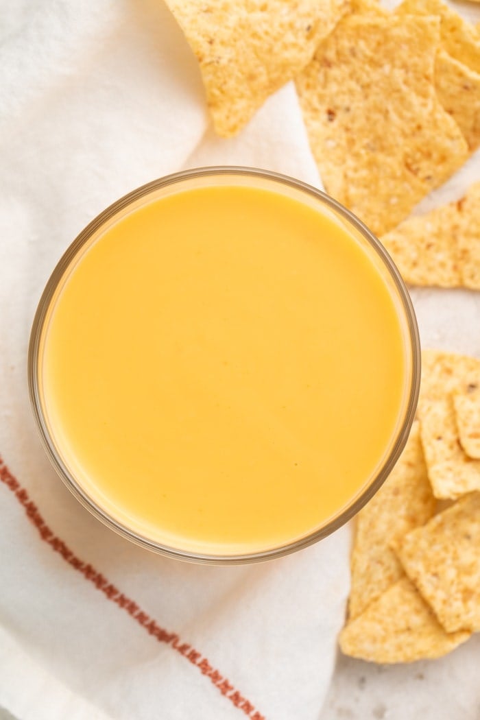 Overhead shot of a clear glass of Taco Bell nacho cheese sauce surrounded by a white napkin with a red line of embroidery and broken tortilla chips