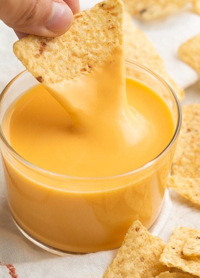 A triangular tortilla chip being dipped into a clear glass of Taco Bell nacho cheese sauce while surrounded by broken tortilla chips
