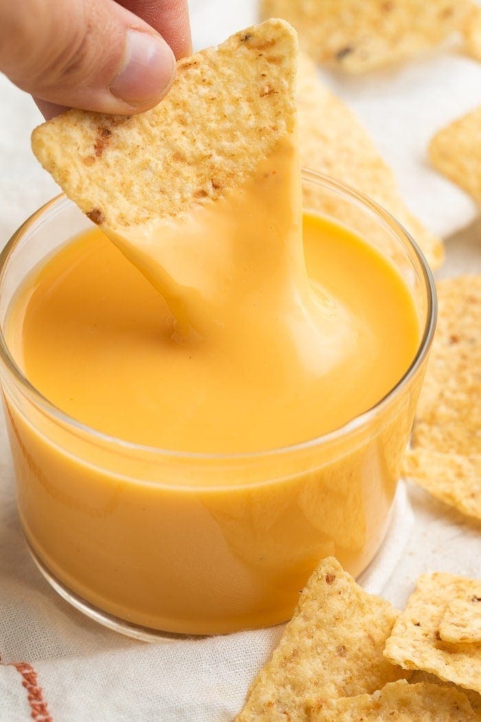 A triangular tortilla chip being dipped into a clear glass of Taco Bell nacho cheese sauce while surrounded by broken tortilla chips