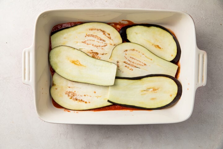 Sliced eggplants layered on top of a bed of bright red tangy marinara sauce in a white casserole dish.