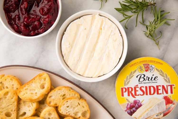 Ingredients for cranberry brie dip on a white tablecloth next to a serving dish of crostini
