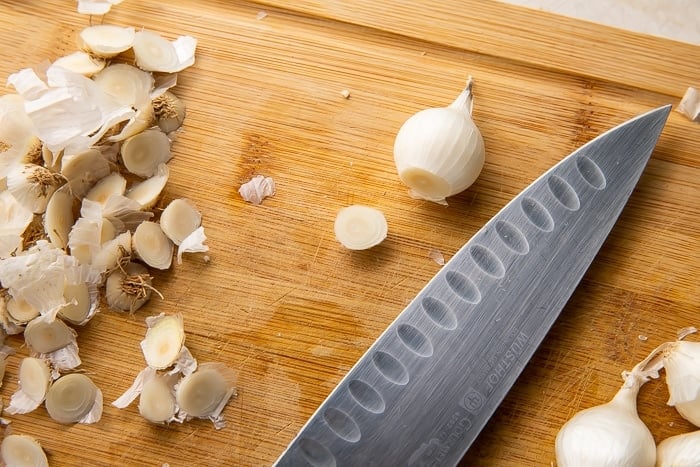 Overhead view of a pearl onion on a cutting board next to a sharp knife. The root end of the onion has been cut away.