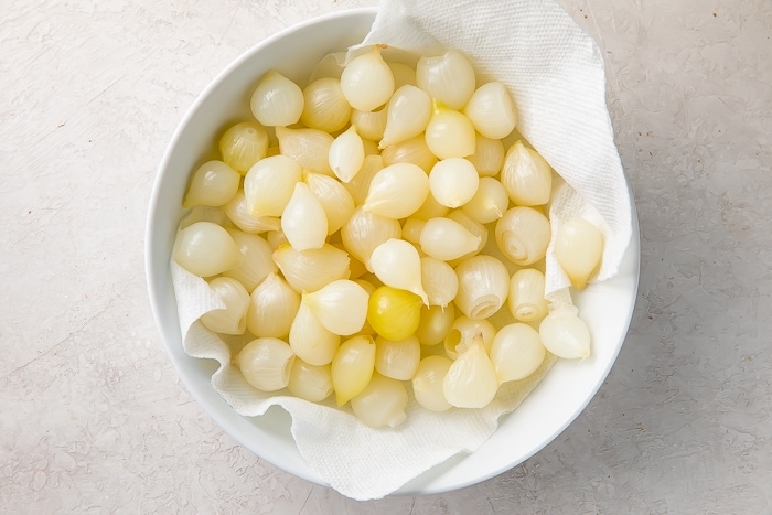 Overhead view of cooked, peeled pearl onions in a large bowl lined with paper towels.