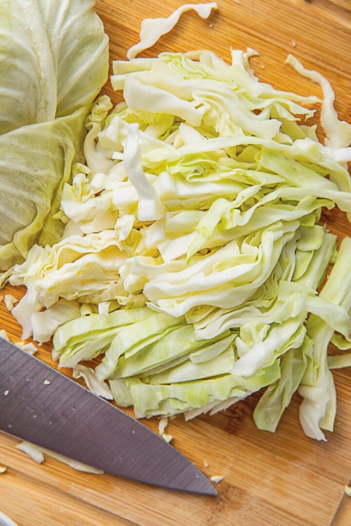 A head of cabbage, shredded, sitting on a wooden cutting board next to a sharp chef's knife.