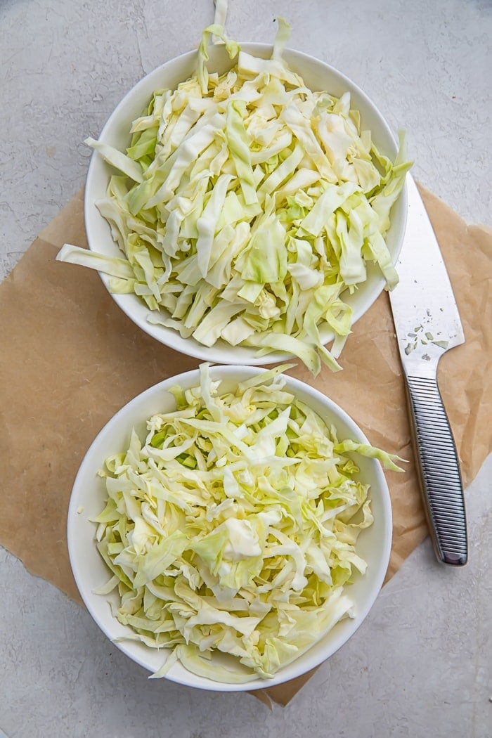 Two bowls of shredded cabbage on a cutting board next to a knife