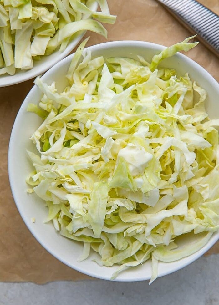 A white bowl full of shredded cabbage on a wooden cutting board next to a knife