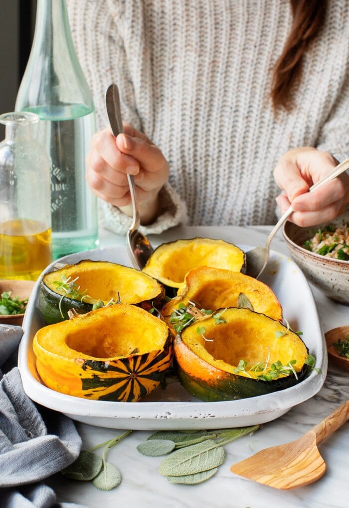 Woman wearing a neutral sweater holding serving utensils removing maple acorn squash from a white serving dish