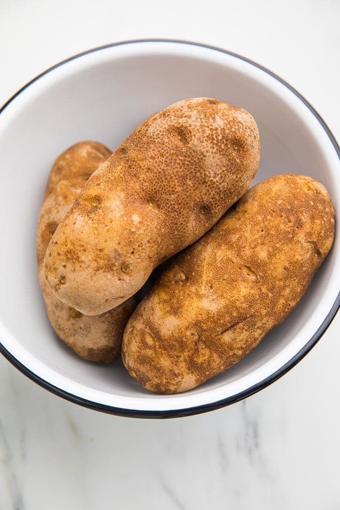 3 unpeeled russet potatoes in a black rimmed white bowl on a marble countertop