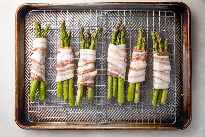 Uncooked asparagus spears wrapped in raw bacon laid perpendicularly on a wire rack over baking sheet