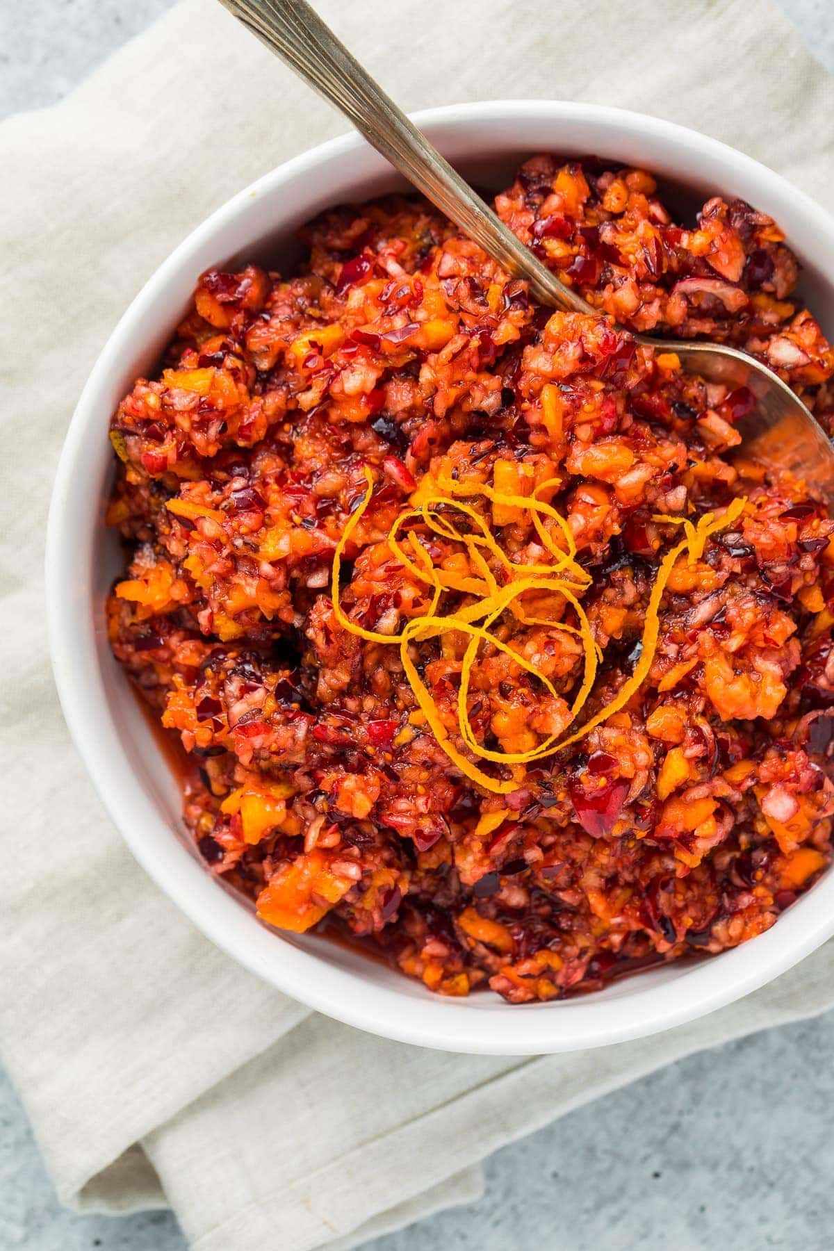 White bowl with cranberry orange relish and garnish and a silver spoon sitting on a white napkin