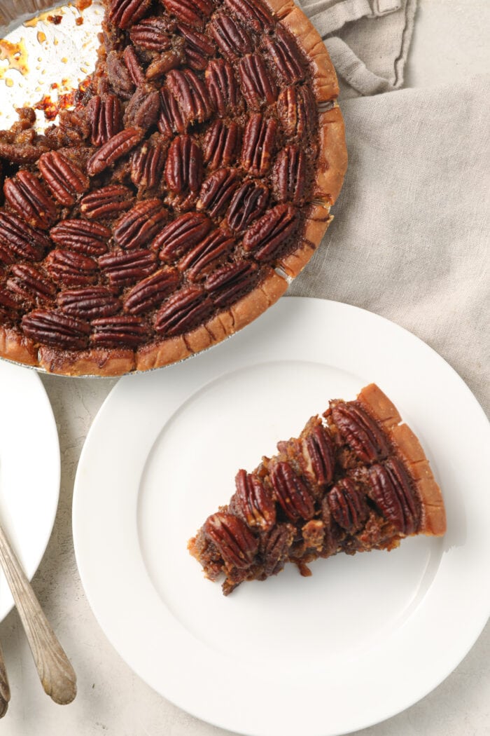 Slice of pecan pie on a white plate next to a whole pecan pie