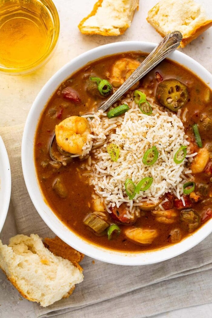 White bowl of shrimp and rice gumbo with a silver spoon surrounded by pieces of bread on a grey napkin with a glass of tea on a white countertop