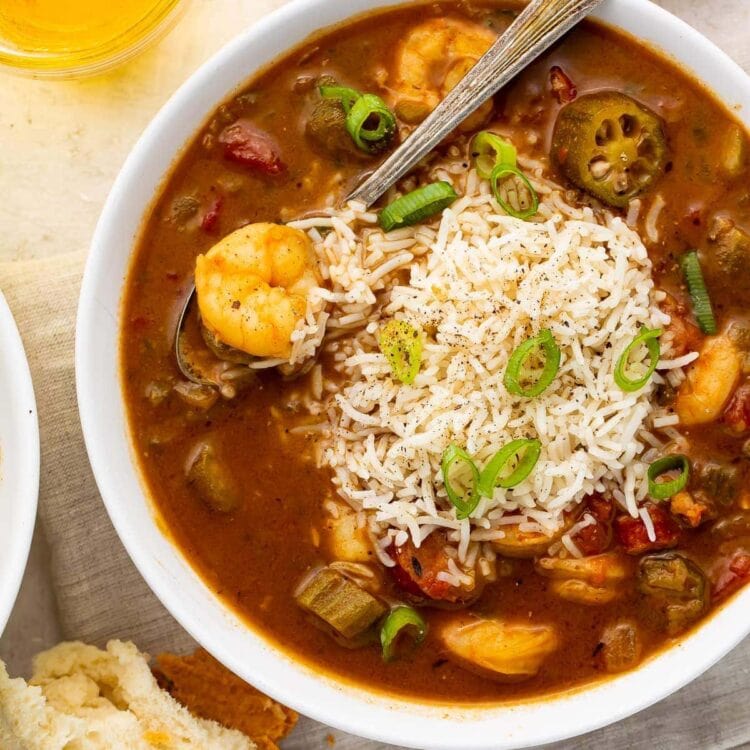 White bowl of shrimp and rice gumbo with a silver spoon surrounded by pieces of bread on a grey napkin with a glass of tea on a white countertop