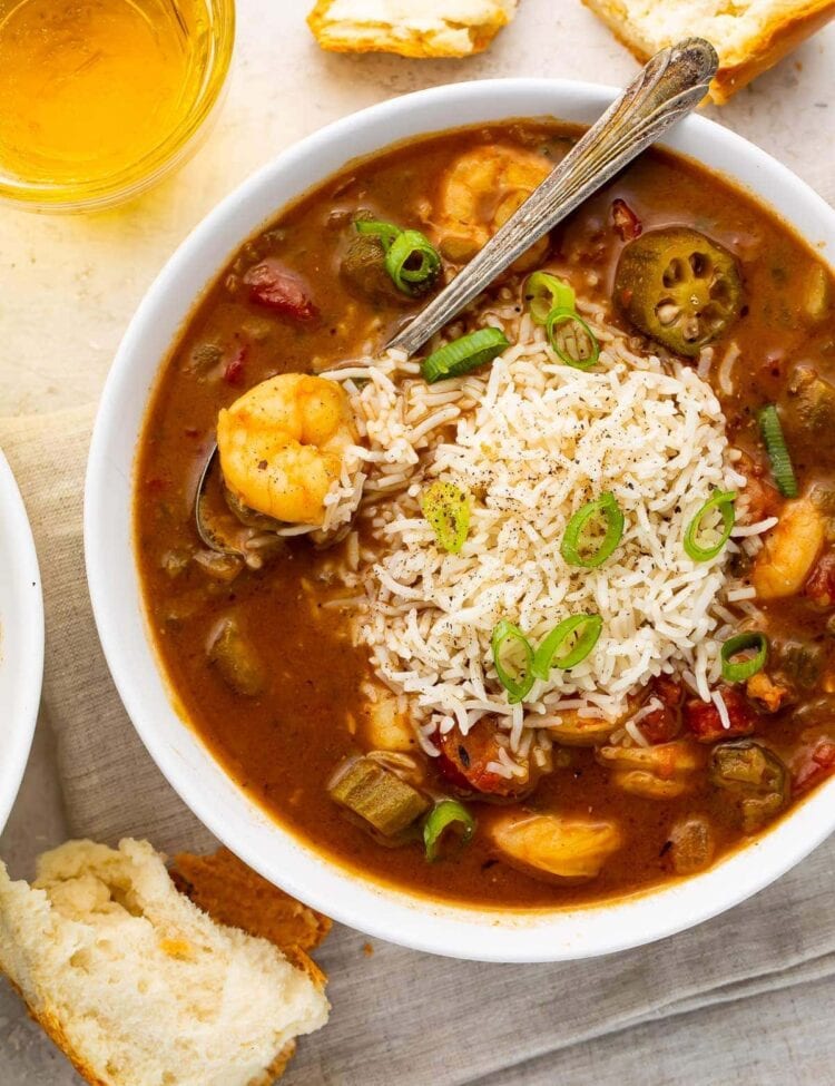 White bowl of shrimp and rice gumbo with a silver spoon surrounded by pieces of bread on a grey napkin with a glass of tea on a white countertop