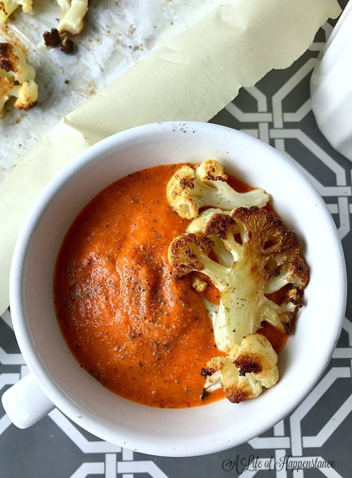 A white bowl of red pepper soup with a roasted cauliflower