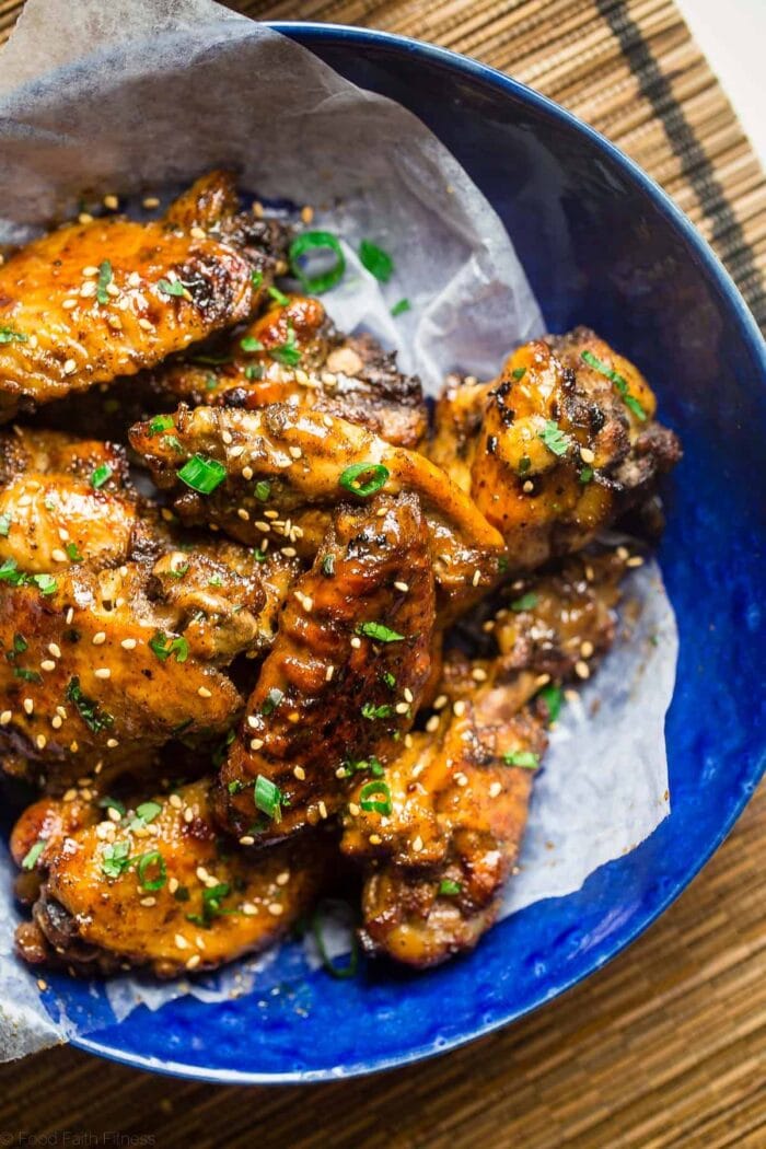 A blue ceramic bowl on a bamboo placemat holds glazed chicken wings with green onions