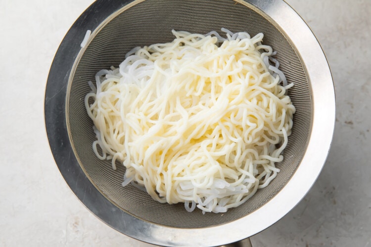 Boiled shirataki noodles in a silver mesh strainer over a silver bowl on a white counter