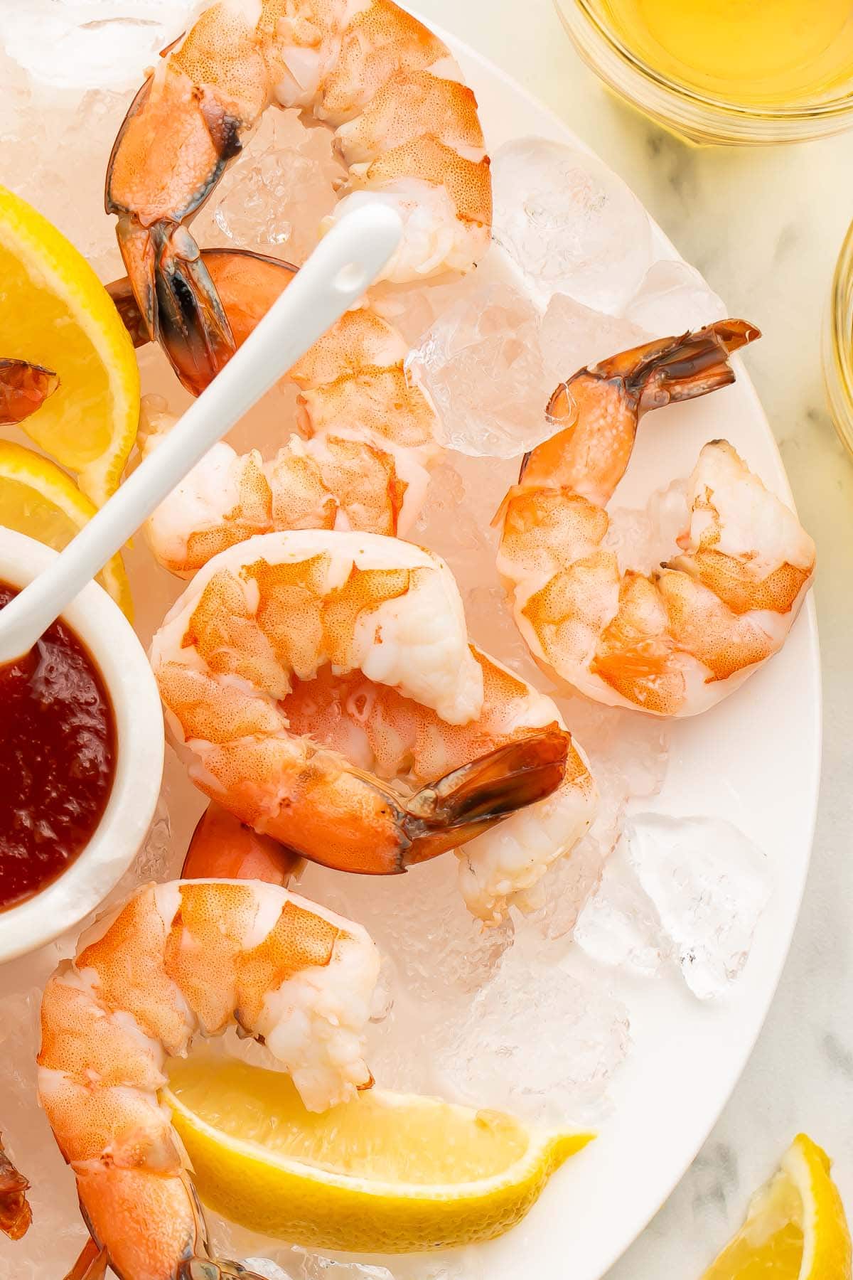 A close-up of poached shrimp arranged on a platter of ice next to a small dipping bowl of shrimp cocktail sauce.