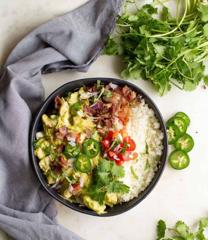 A black bowl containing jalapenos, chicken, and cauliflower rice on a white table surrounded by jalapenos