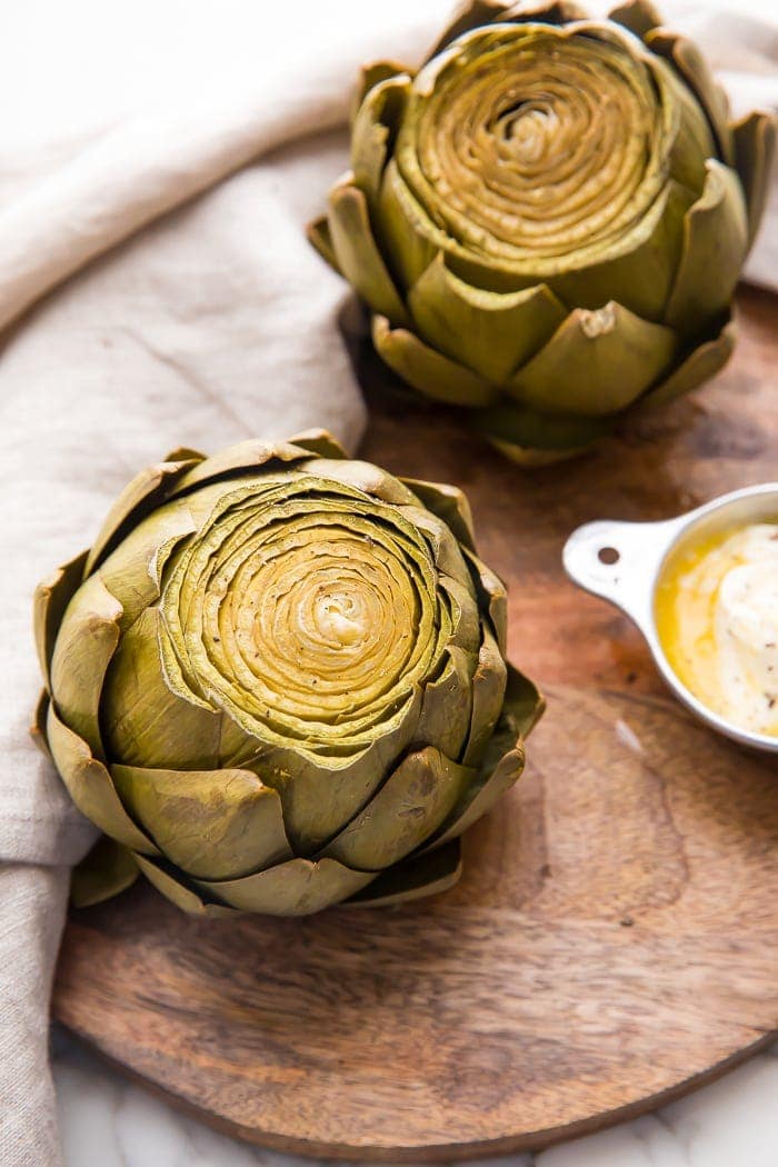 Two Instant Pot cooked artichokes on a wooden cutting tray with dipping sauce and a napkin