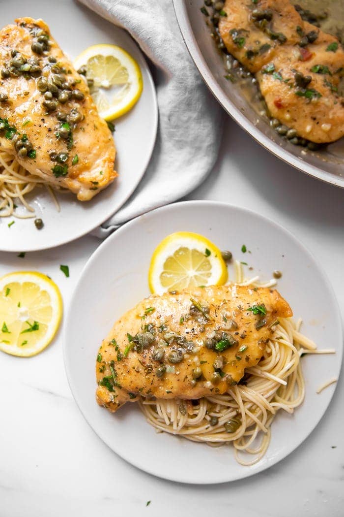 Chicken piccata on top of veggie noodles with a slice of lemon on a white plate