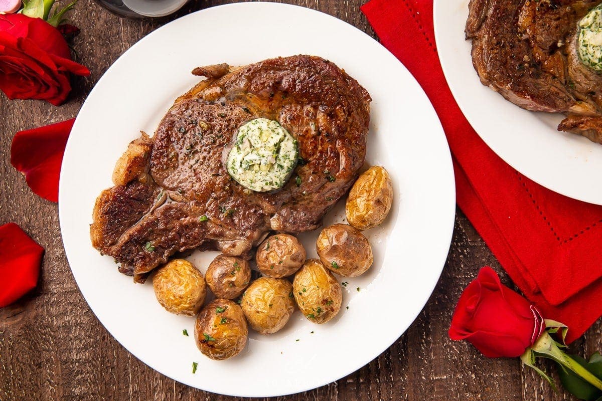 Whole bone-in ribeye with garlic-herb butter on a white plate with whole roasted potatoes surrounded by red roses and a red napkin on a wooden table