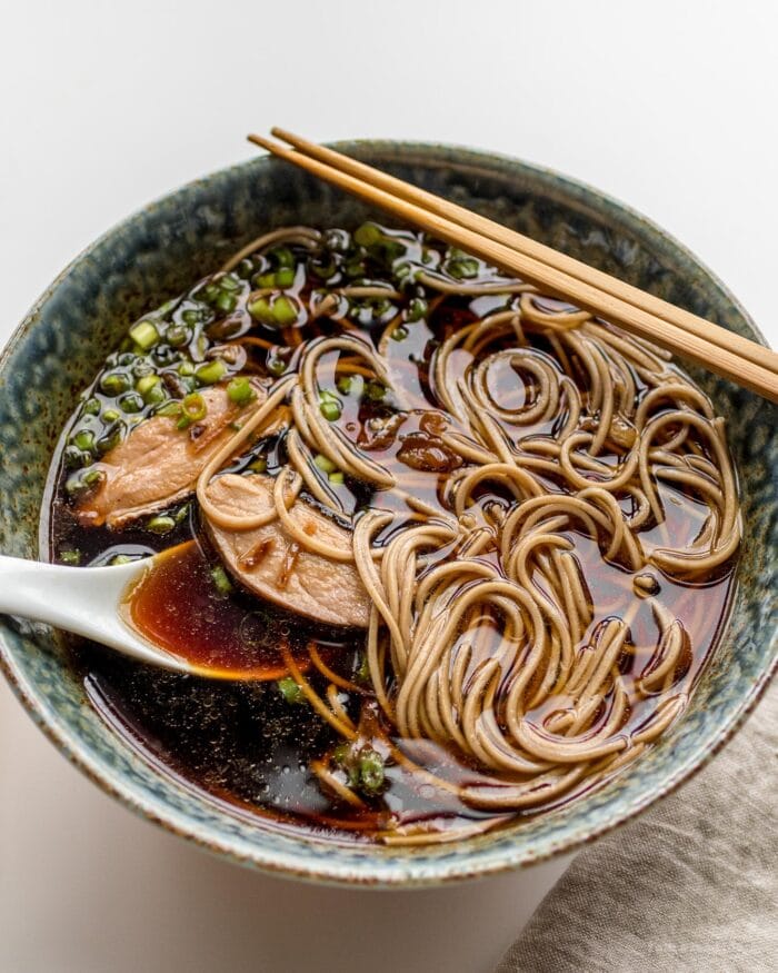 A bowl of ramen, duck, and broth, with a spoon and chopsticks