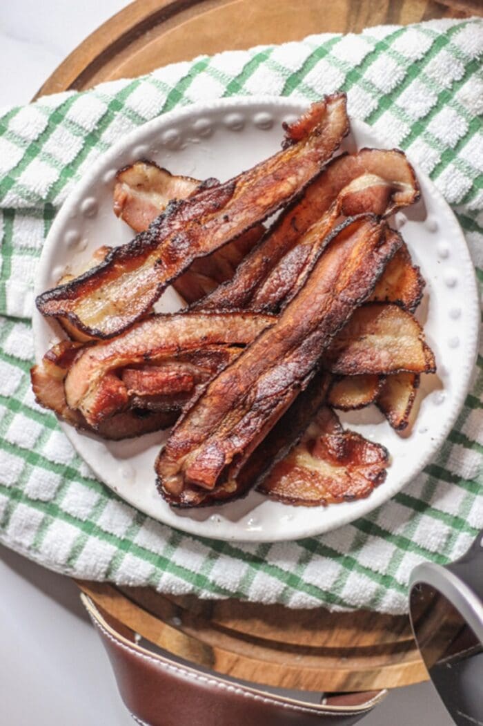 A plate of bacon on top of a green and white checkered dish towel on a wooden cutting board