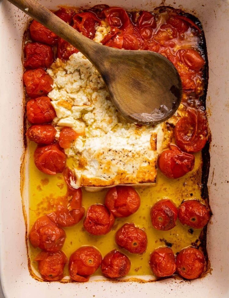 Baked feta and tomatoes smushed in oil in a large pan with a wooden spoon
