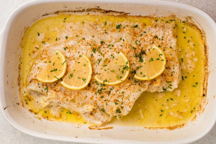 Baked fish topped with parsley and lemon slices in a white baking dish