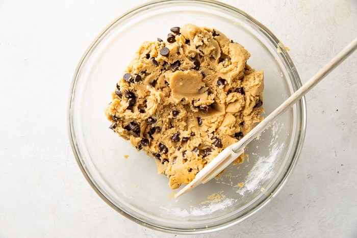 Vegan chocolate chips added to dough in a glass mixing bowl with a spatula
