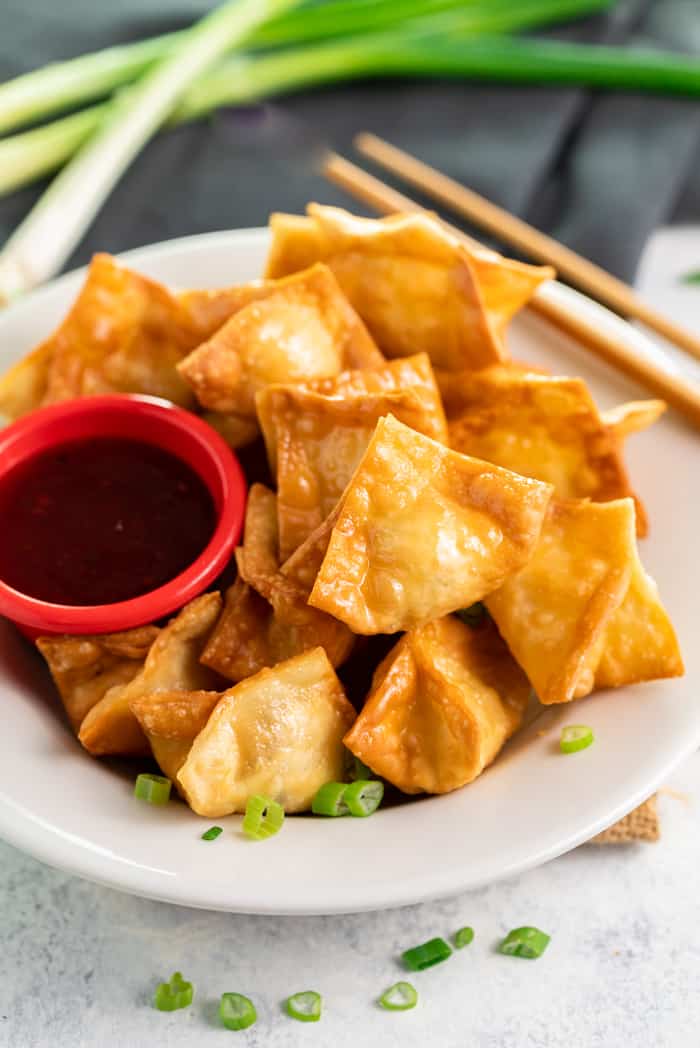 A white plate of crab rangoon with chopsticks and a small red dipping bowl filled with sauce